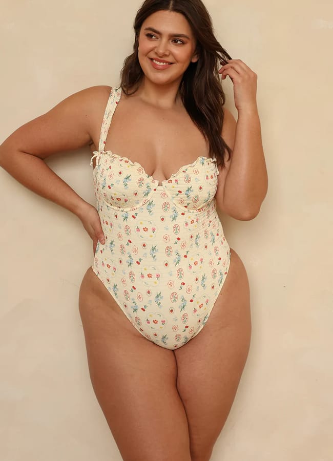A woman smiles while wearing pretty swimwear—a floral one-piece swimming costume with a sweetheart neckline and adjustable straps—standing against a beige background.