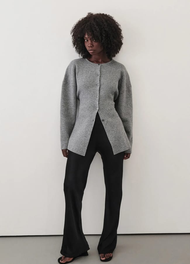 A woman with curly hair stands against a plain wall, wearing a grey button-up cardigan and black trousers, looking slightly to the side with a neutral expression—perfect for updating your wardrobe during the summer sale.