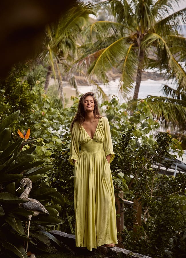 A woman in a long, light green dress stands outdoors among lush tropical plants and palm trees, with the sea and waves visible in the background—capturing the carefree spirit of a last minute holiday.