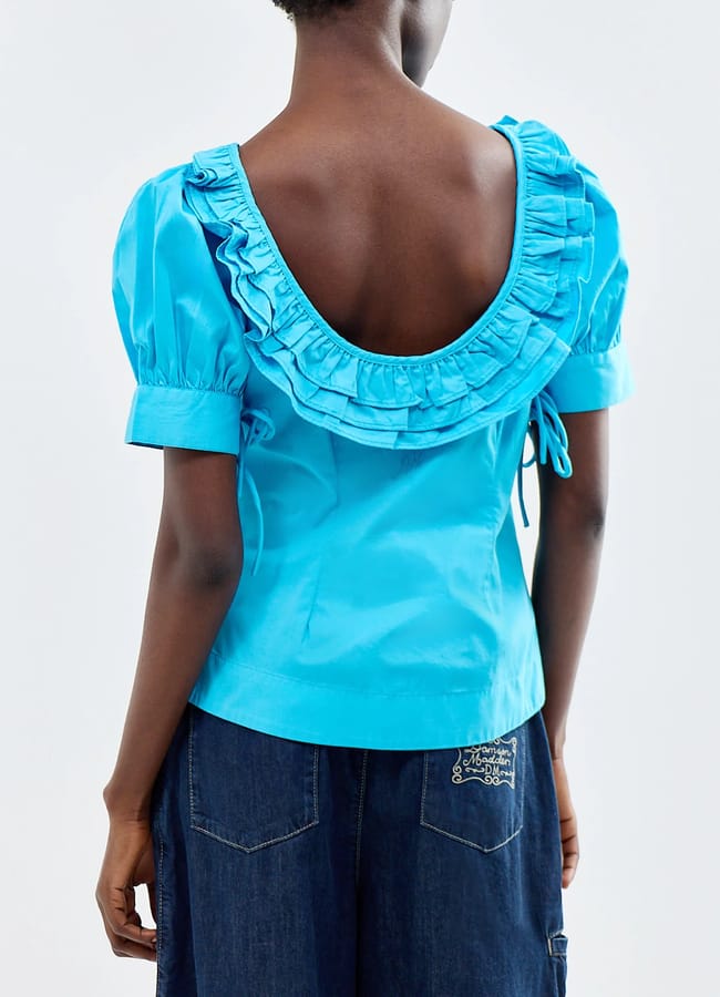 A person wearing a bright blue, short-sleeve blouse inspired by turquoise fashion, featuring a deep ruffled scoop back and puffed sleeves, paired with dark denim trousers. The photo is taken from behind.
