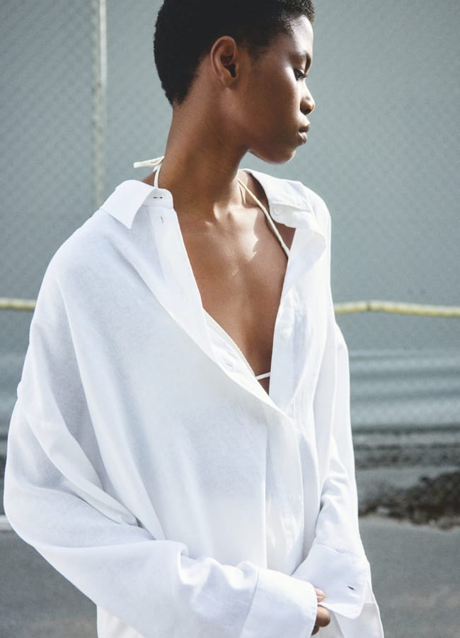 A person with short hair stands sideways, wearing an oversized white button-up shirt over a white top, outdoors near a chain-link fence—capturing the relaxed vibe of a last minute holiday.