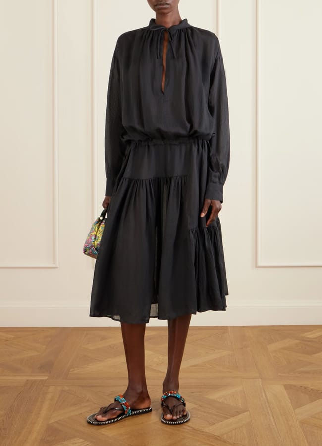 A person wearing a loose-fitting, long-sleeved black dress, colourful sandals, and holding a multicoloured bag stands on a wooden floor against a cream-panelled wall—an effortlessly chic look perfect for the summer sale season.