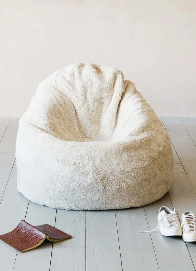 A soft, cream-coloured bean bag chair sits on a light wooden floor—perfect for relaxing during a night in watching a film. In front of it are a pair of white trainers and an open brown notebook. The background is a plain, pale wall.