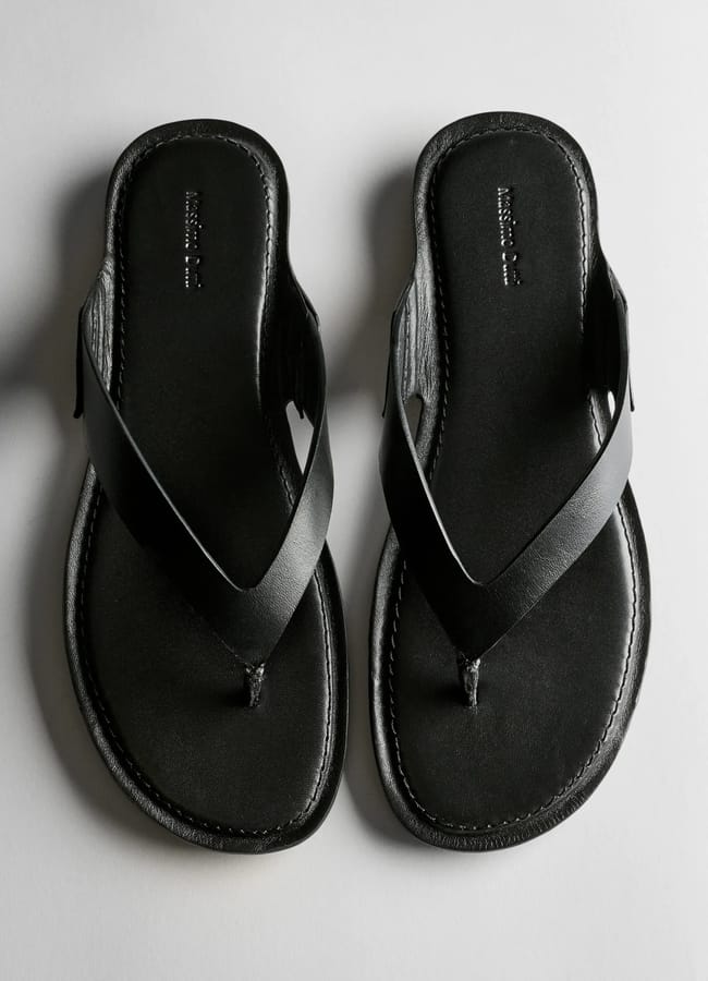 A pair of black leather thong sandals with stitched edges, photographed from above on a light grey background—perfect for complementing your favourite oversized shirt outfit ideas.