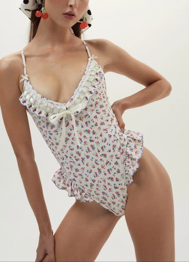 Woman wearing a white floral ruffled swimming costume with a ribbon tie at the chest, showcasing pretty swimwear against a plain light background. She has statement earrings and a spotted hair accessory.