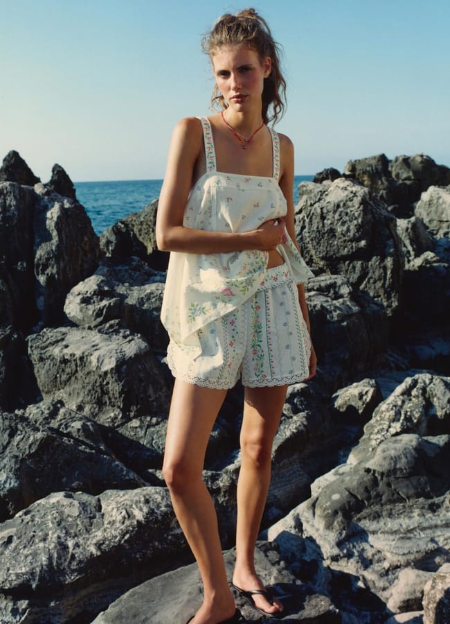 A woman stands on rocky terrain by the sea, wearing a light floral top and matching shorts, with flip-flops—perfect attire for a last minute holiday. The sky is clear and blue, and she looks at the camera with a relaxed expression.