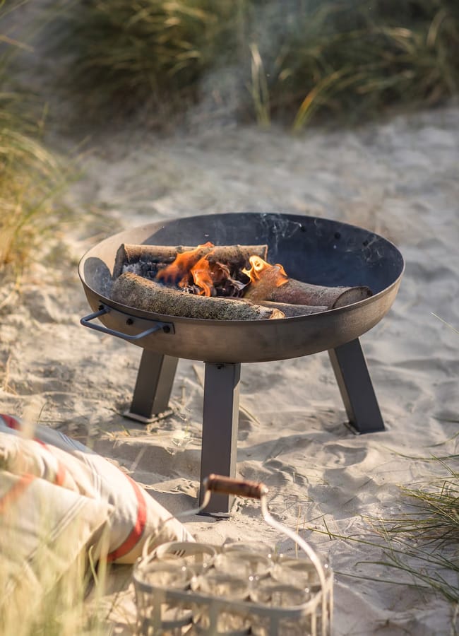 A black metal firepit with burning logs sits on sandy ground surrounded by beach grass. A striped cushion and a lantern are nearby, creating a cosy outdoor setting—perfect for an outdoor film night under the stars.