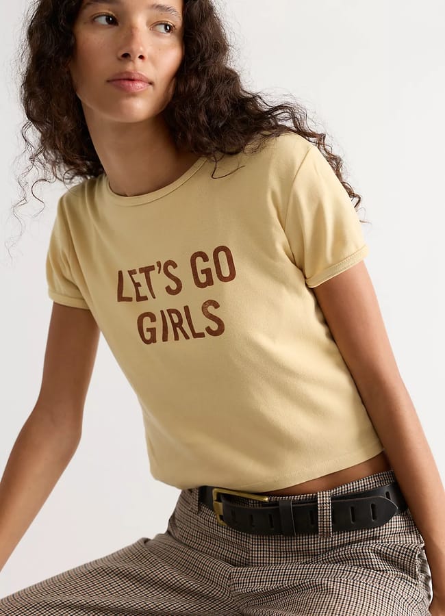A woman with long curly hair wears a yellow T-shirt—one of this season’s cool slogan tees—that says LET'S GO GIRLS in brown letters and checked trousers with a black belt, sitting casually and looking to the side.