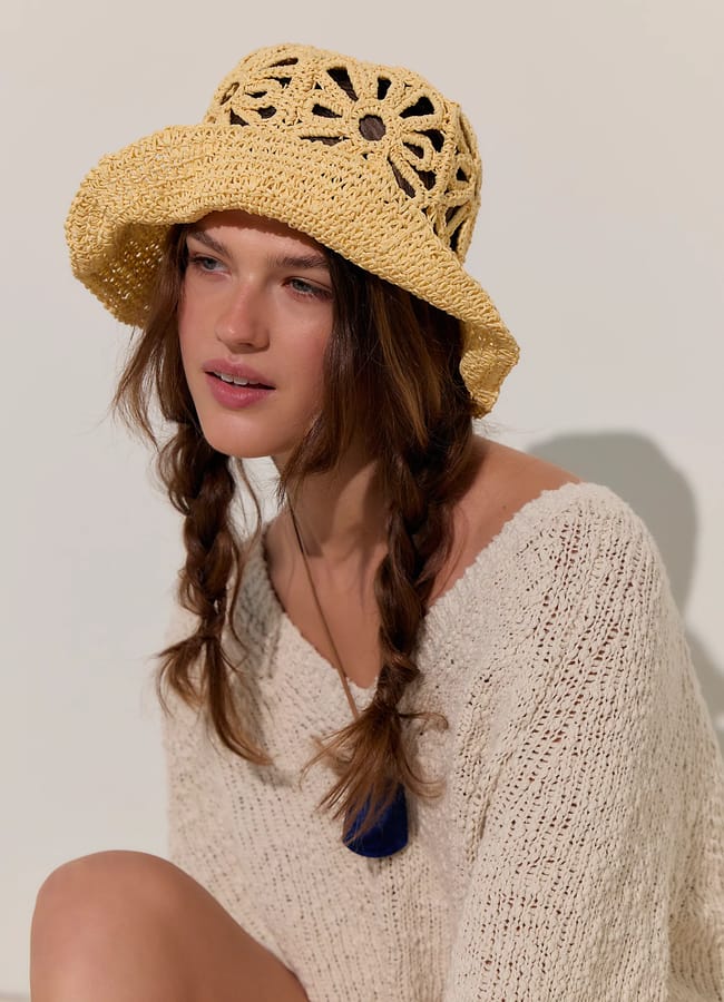 A woman with brown plaited hair wears a yellow crochet sun hat and a loose-knit cream jumper, sitting against a neutral background—ready for that effortless last-minute holiday escape.