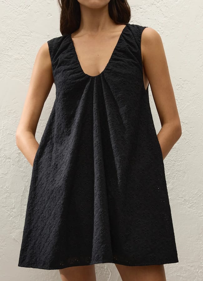 A woman wearing a sleeveless, loose-fitting black dress with a deep V-neckline and subtle broderie anglaise pattern stands against a light textured wall, with her hands in the dress pockets—perfect for those warm days and now available during our summer sale.