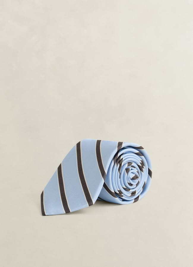 A light blue necktie with dark diagonal stripes, neatly rolled on a plain beige surface—a perfect touch of men’s summer dressing inspired by The Talented Mr Ripley style.