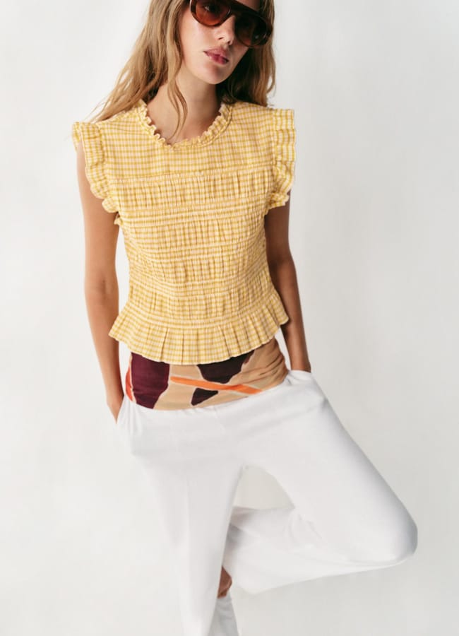 Woman wearing oversized sunglasses, a yellow ruffled sleeveless top with a chequered pattern, and white trousers, standing against a plain white background with hands in her pockets—perfect inspiration for your next holiday fashion look or those spontaneous last minute holiday pics.