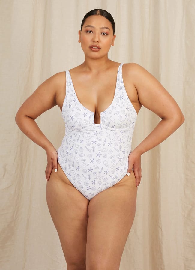 A woman stands confidently in pretty swimwear—a white one-piece swimming costume with a subtle leaf pattern, hands on hips, against a beige fabric backdrop.