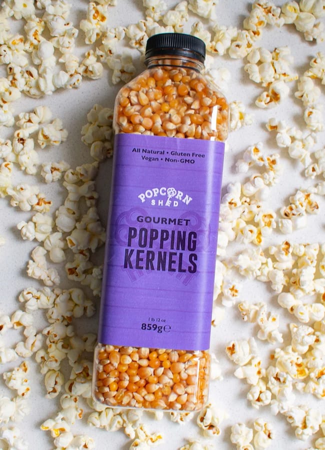 A bottle of Popcorn Shed Gourmet Popping Kernels lies on a white surface surrounded by scattered popped popcorn, ready to elevate your next outdoor film night at home. The bottle features a purple label with white and black text, promising delicious popcorn for any cosy evening under the stars.