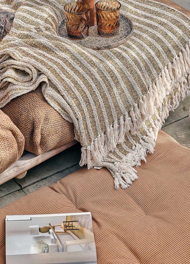 A cosy scene reminiscent of an outdoor-film-night-at-home, featuring a brown textured cushion, a striped fringed throw, two amber glasses on a tray, a grey dog, a ribbed lamp, and an open magazine on a matching floor cushion.