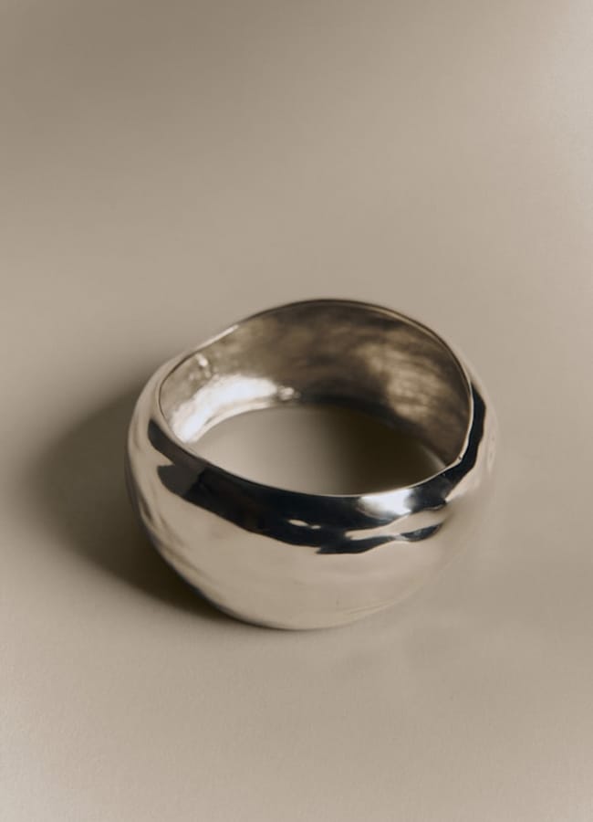 A smooth, shiny, silver bangle with an irregular, organic shape sits on a neutral beige surface. The bracelet’s surface reflects light, giving it a modern and minimalist appearance—perfect for pairing with your favourite oversized shirt outfit ideas for an effortlessly chic look.