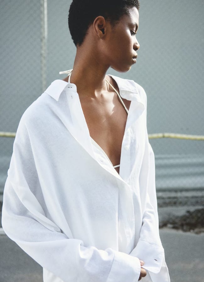 A person with short hair wearing an oversized white shirt stands outdoors, embodying minimalist summer fashion. They look down and to the side, with a chain-link fence and blurred background visible behind them.