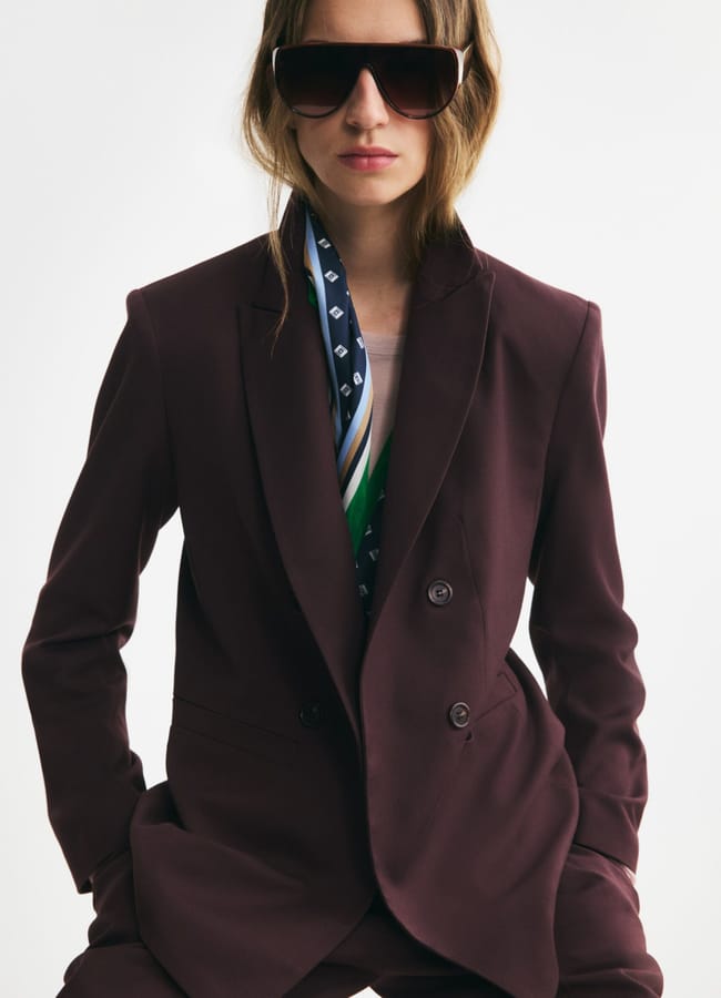 A person in a burgundy suit, patterned scarf, and large dark sunglasses poses confidently against a plain background, effortlessly embodying the bold elegance of autumn winter 2026 fashion trends.