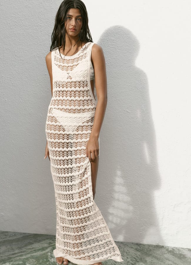A woman with long dark hair stands barefoot on a green stone floor, wearing a sleeveless, sheer white lace dress over a white bikini—perfect for a last minute holiday escape. She stands against a white textured wall, her shadow cast beside her.