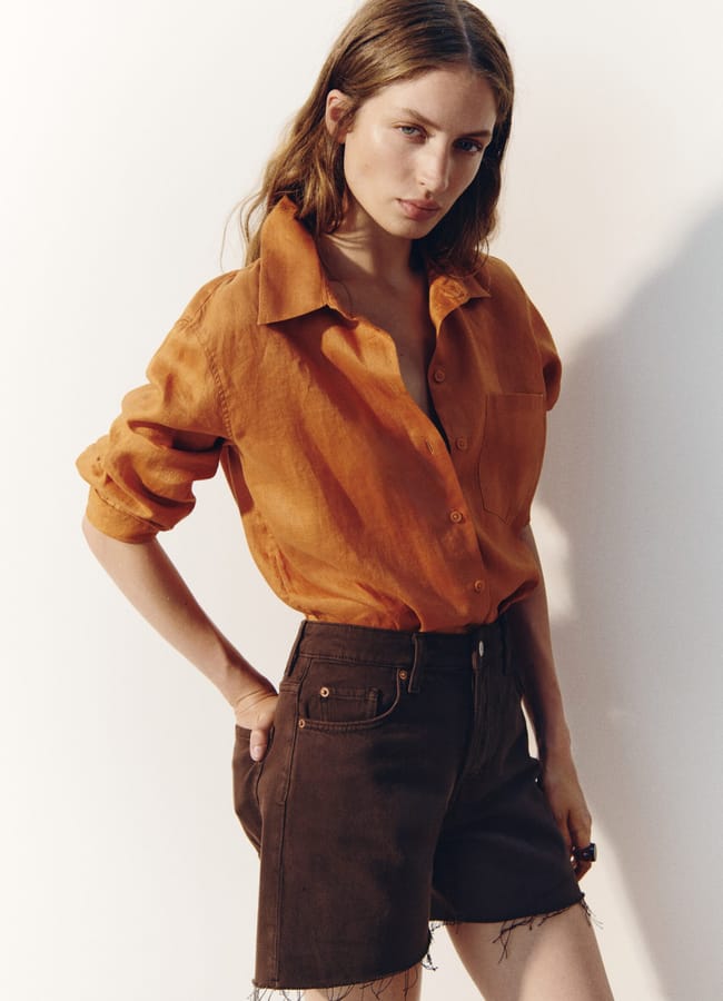 A woman stands against a light background wearing an orange button-up shirt with rolled sleeves and dark brown cut-off shorts, her left hand in her pocket—ready for a spontaneous last-minute holiday adventure.
