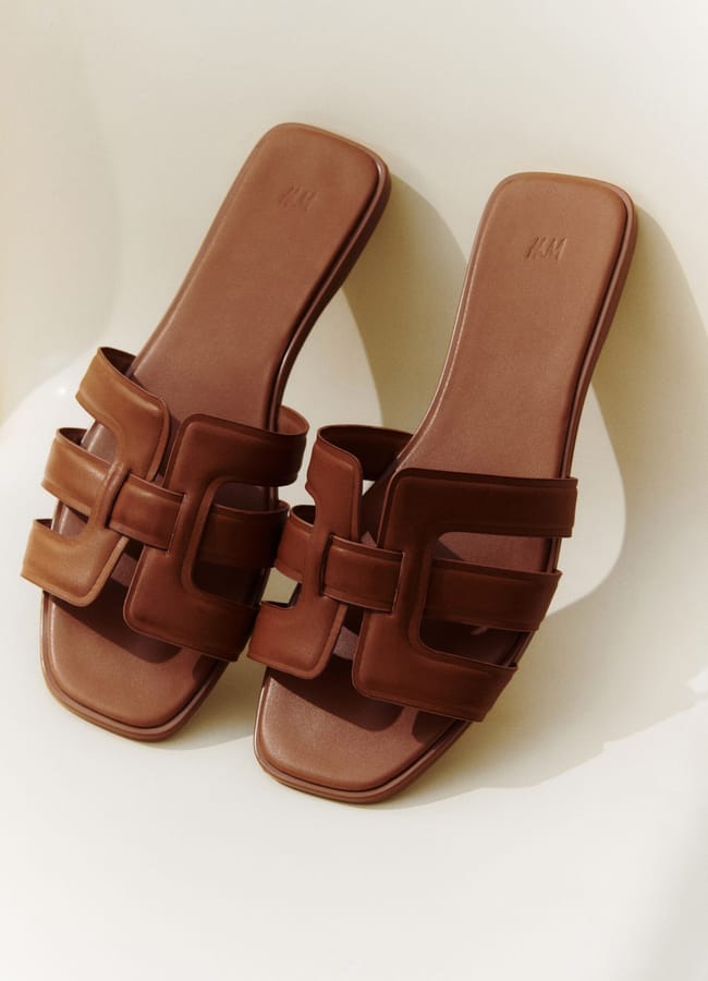 A pair of brown open-toe slide sandals with cut-out geometric straps, placed side by side on a light-coloured surface—perfect for slipping on during your last-minute holiday getaway.