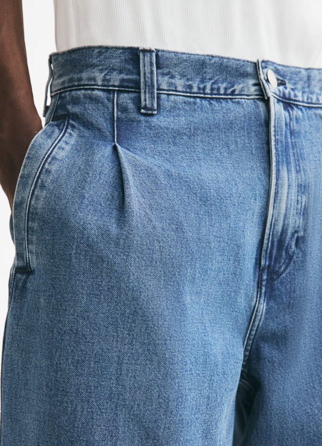 Close-up of a person wearing light blue pleated denim jeans and a white ribbed top, with one hand in the pocket. The jeans feature belt loops and a visible metal button at the waistband, perfectly styled for a smart-casual look when paired with a men’s blazer and jeans ensemble.