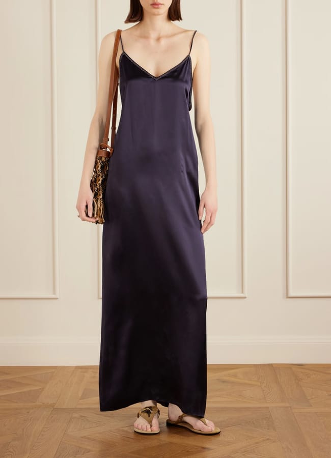 A woman stands against a cream wall, wearing a dark navy satin slip dress perfect for the summer sale, a brown shoulder bag with tassels, and flat sandals. The dress has thin straps and a V-neckline. The setting has a wooden floor and white panelling.