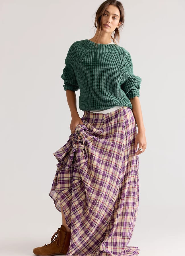 A woman poses wearing a chunky green jumper and a long purple check skirt, holding up one side. Reflecting autumn winter 2026 fashion trends, she pairs the look with brown ankle boots and loose hair, standing against a plain light background.