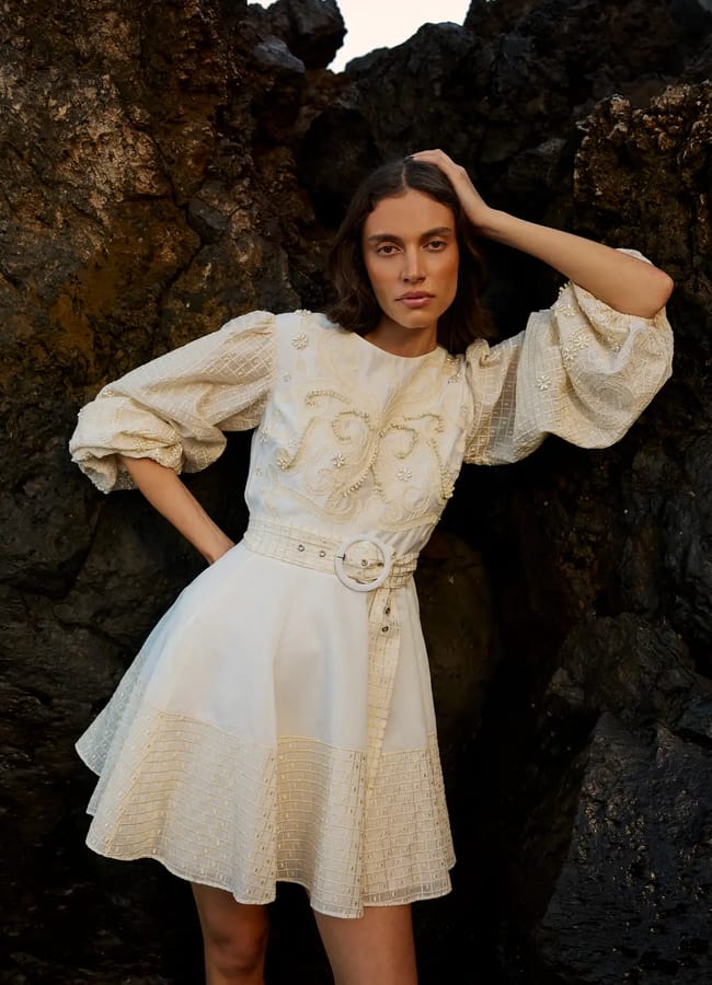 A woman in a white, long-sleeved belted dress stands against dark rocky cliffs, resting one hand on her head and gazing at the camera—a timeless choice for effortless holiday style.