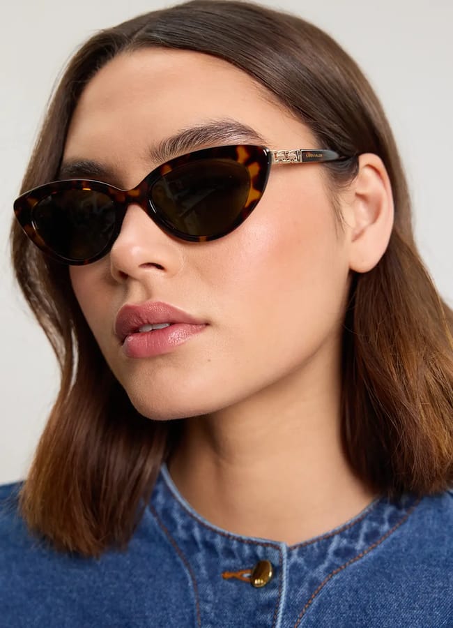 Woman with straight brown hair wearing cat-eye tortoiseshell sunglasses and a blue denim top, looking slightly to the side against a plain background—perfect inspiration for your holiday fashion or last minute holiday snaps.