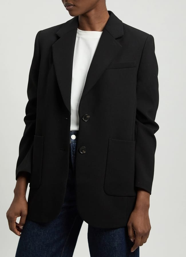 Person wearing a black blazer over a white t-shirt and dark blue jeans, standing against a plain background. This look captures the essence of minimalist summer fashion, with the blazer featuring two front pockets and fastened with two buttons. The person’s head is partially out of frame.