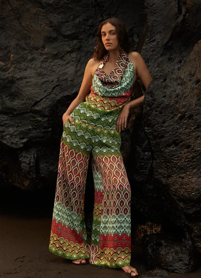 Woman stands against a dark rock wall, wearing a colourful, patterned halter-neck jumpsuit with flared legs and sandals—a perfect holiday fashion choice for capturing memorable holiday photos. She has loose, wavy hair and a relaxed pose, gazing at the camera.