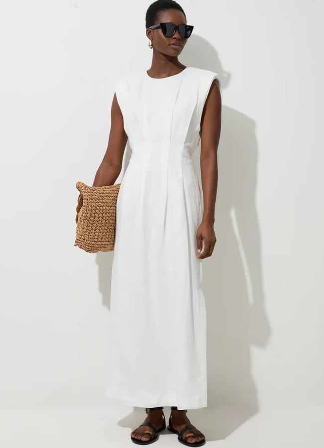 Person wearing a sleeveless white maxi dress, dark sunglasses, and brown sandals holds a woven straw bag whilst standing against a plain white background—perfect inspiration for last minute holiday fashion or creating effortless holiday fashion pics.