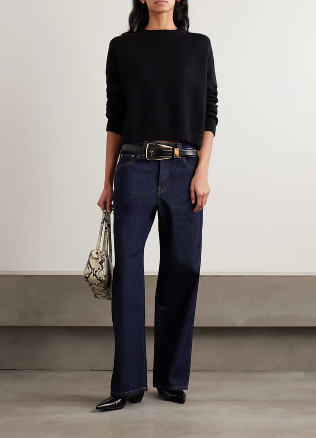 A person wearing a black jumper, dark wide-leg jeans with a belt, black pointed shoes, and holding a small snake-print handbag stands indoors against a plain wall and concrete floor—an effortlessly chic look perfect for discovering new favourites during the summer sale.