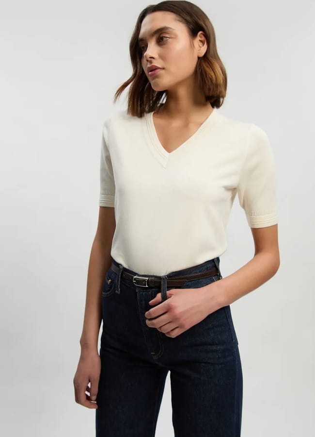 A woman with shoulder-length brown hair wears a short-sleeved, cream V-neck jumper tucked into dark blue jeans with a black belt, effortlessly showcasing the coolest way to embrace everyday fashion as she stands against a plain light background.