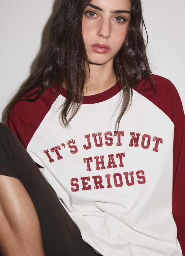 Young woman with long dark hair wearing one of those cool slogan tees—a white and red raglan shirt that says “It’s just not that serious.” She sits against a plain white background, looking directly at the camera with a neutral expression.