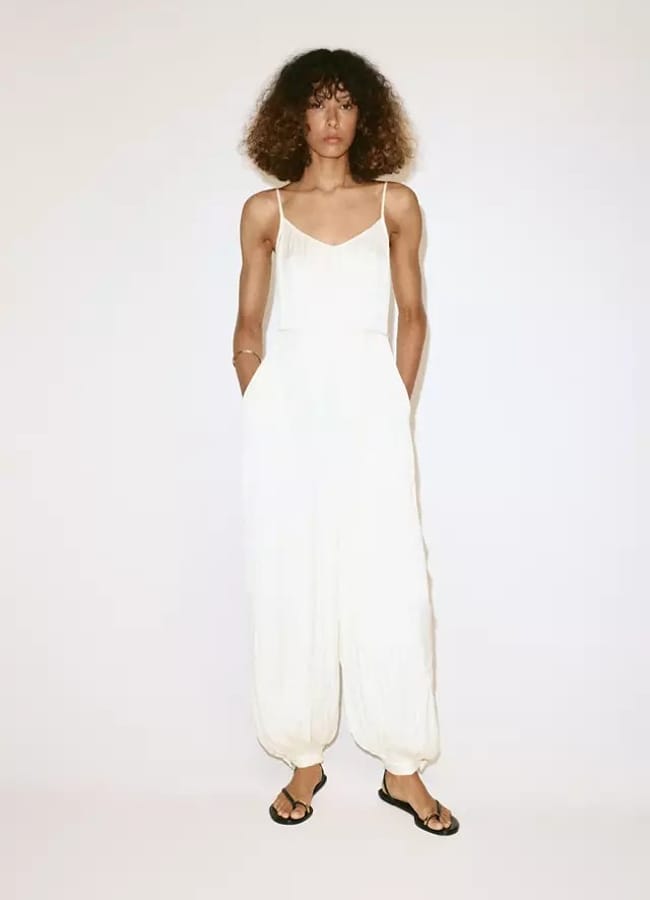 A person with curly hair wears a loose white playsuit with spaghetti straps and elasticated cuffs, paired with black sandals, standing against a plain light background—an effortlessly chic choice for last-minute holiday outfits.