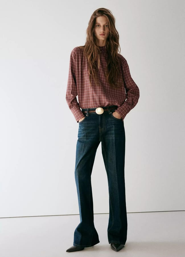 Person wearing a red checked shirt tucked into high-waisted blue jeans with a belt, standing against a plain white background with their hands in their pockets—an effortlessly stylish look that perfectly embodies autumn winter 2026 fashion trends.