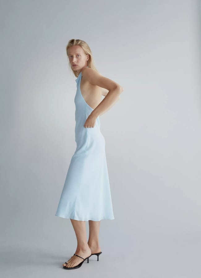 A woman with long blonde hair wearing a light blue, sleeveless, backless midi dress and black heeled sandals stands sideways against a plain light grey background, perfectly styled for that effortless last minute holiday getaway.