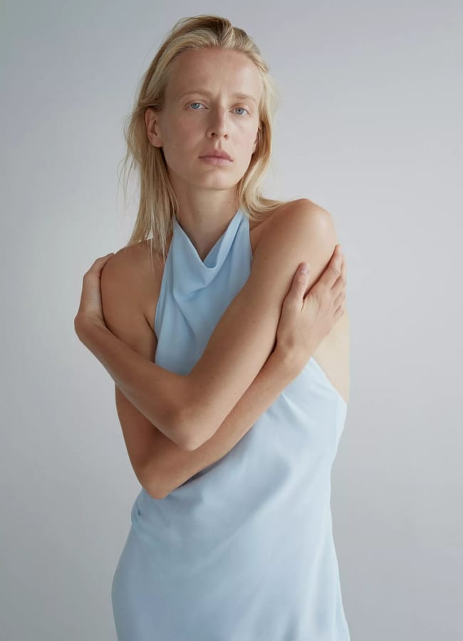 A person with long blonde hair wears a light blue halter dress and crosses their arms over their chest, standing against a plain light background—capturing the relaxed vibe of a last minute holiday.