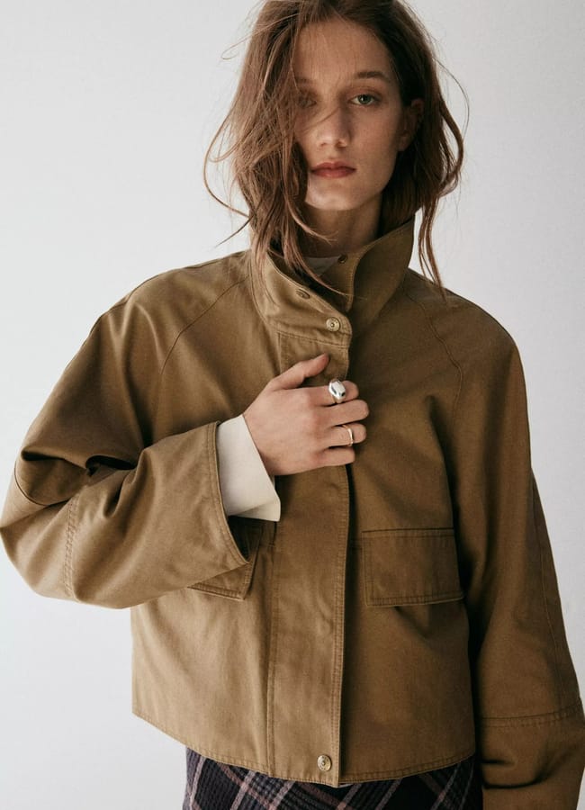 A woman with tousled hair wearing a brown jacket over a light top and checked trousers stands against a plain background, looking towards the camera with a neutral expression—an effortlessly chic look that captures the essence of autumn winter 2026 fashion trends.