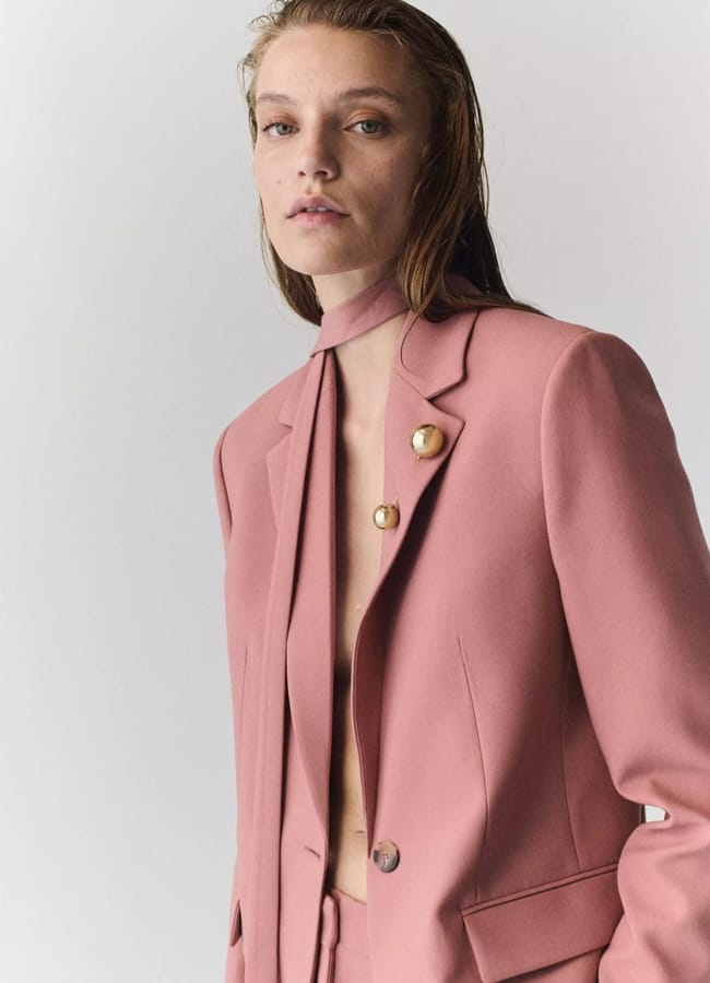A person with wet hair wears a dusty pink suit jacket with gold buttons, no shirt underneath, and a matching necktie, standing against a light grey background—a striking look inspired by autumn winter 2026 fashion trends.