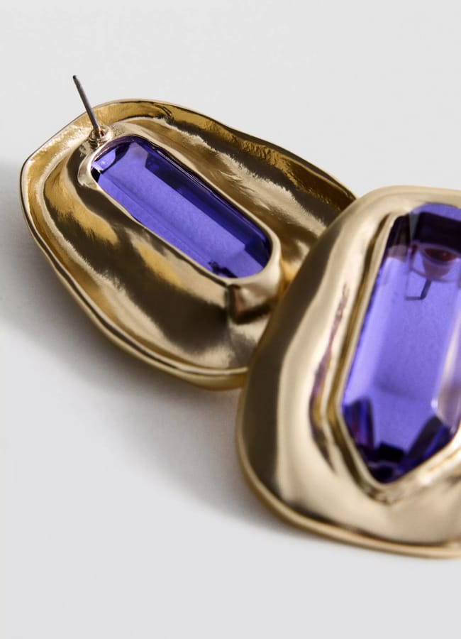 Close-up of two gold earrings with large purple gemstones set in a textured, irregular gold frame, placed on a white surface—a striking accessory that captures the bold elegance seen in autumn winter 2026 fashion trends.