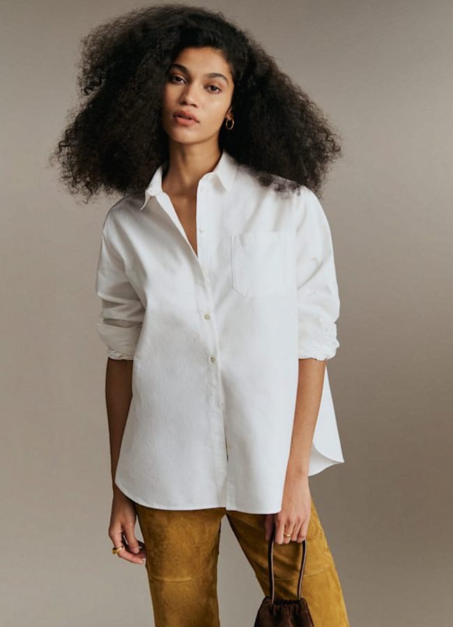 Woman with voluminous curly hair wears a white oversized shirt, offering fresh inspiration for oversized shirt outfit ideas, paired effortlessly with mustard yellow trousers and a small brown handbag, standing against a neutral background.