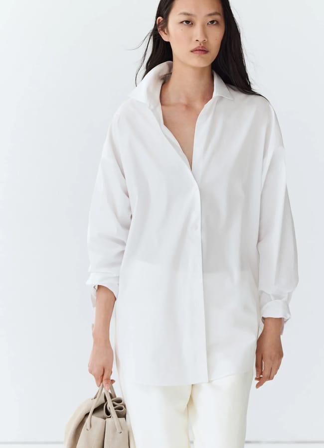 A woman with long dark hair wears a loose white button-up shirt and white trousers, holding a beige bag against a plain white background—an effortlessly chic example for those seeking oversized shirt outfit ideas.