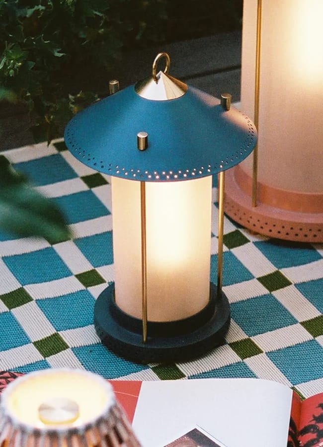 A lantern with a blue top and gold accents sits on a chequered tablecloth, glowing softly—perfect for setting the mood at your outdoor film night. Nearby are a pink lantern, some greenery, and an open book with colourful pages, creating a cosy atmosphere to enjoy your at-home film night under the stars.