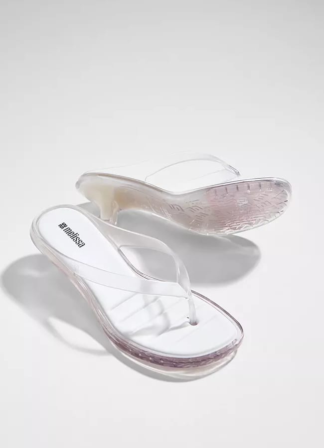 A pair of clear wedge flip-flop sandals with white footbeds, displayed on a light background. Reminiscent of the chic style often spotted on celebrity couples on holiday, one sandal is upright while the other is tilted to show the translucent sole.