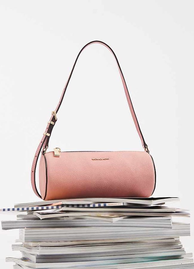 A pink cylindrical handbag with a shoulder strap sits atop a stack of magazines—perhaps the latest issues featuring Celebrity Couples On Holiday—against a plain white background.