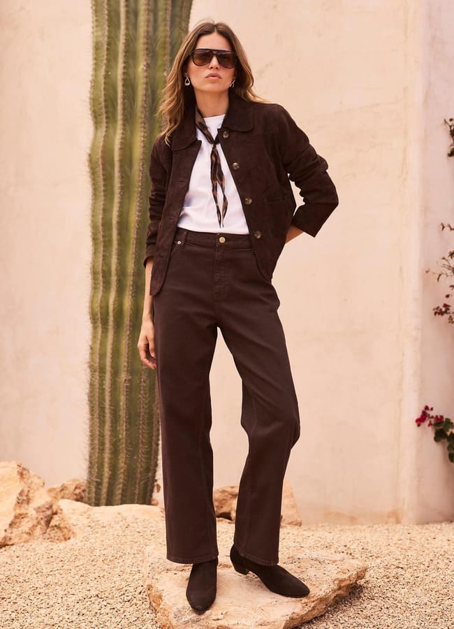 A woman wearing dark brown trousers, a matching jacket, white shirt, scarf, sunglasses, and black boots stands confidently outdoors near a large cactus and beige wall, showcasing one of the standout autumn winter 2026 fashion trends.