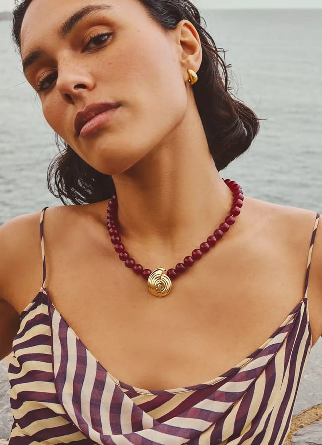 A woman wearing a striped dress, stylish fashion accessories including a red beaded necklace with a gold pendant and gold hoop earrings, poses outdoors near water, looking confidently at the camera.